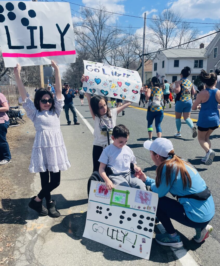 Lily kneeling down to her student in a wheelchair who is holding a sign that says "Go Lily" in English and Braille. 2 other students are standing next to them holding similar signs.