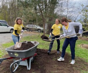 3 Corporate Group Volunteers from Liberty Mutual wearing yellow company shirts are smiling while filling a wheelbarrow with dirt on The Carroll Center for the Blind campus.
