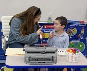TVI and student working A TVI and student are sitting at a table together. The TVI is giving instructions to student. There is a Brailler and markers on the table.