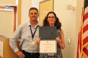 Jill Suarez and Carroll Center President & CEO, Greg Donnelly, stand together smiling in a brightly lit room with the American flag visible to the right. Jill is holding a blue certificate folder with an award. The text on the certificate reads: "The Bill McMahon Employee of Outstanding Distinction Award is presented to Jill Suarez." 