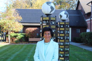 Carol Covell, a woman in a light blue jacket is standing on The Carroll Center for the Blind campus. Behind her are two tall structures with globes on top and rows of small bells. The structures have plaques with inscriptions. The setting appears to be a garden or outdoor area with trees, bushes, and a bench. The background includes a building with a dark roof.