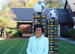 Carol Covell, a woman in a light blue jacket is standing on a well-manicured lawn. Behind her are two tall structures with globes on top and rows of small bells. The structures have plaques with inscriptions. The setting appears to be a garden or outdoor area with trees, bushes, and a bench. The background includes a building with a dark roof.