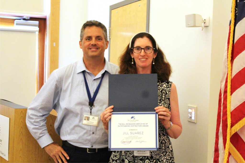 Jill Suarez and Carroll Center President & CEO, Greg Donnelly, stand together smiling in a brightly lit room with the American flag visible to the right. Jill is holding a blue certificate folder with an award. The text on the certificate reads: "The Bill McMahon Employee of Outstanding Distinction Award is presented to Jill Suarez.