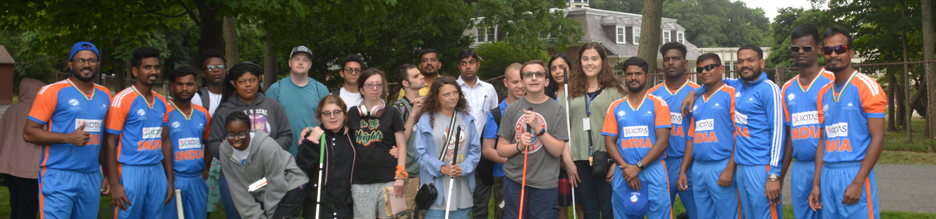 A group of people, including individuals who are blind or visually impaired, poses together outdoors on a grassy area surrounded by trees. Some individuals are holding white canes. Among them, a cricket team dressed in blue and orange uniforms with 'India' written on their jerseys is also present, with some players holding cricket bats. The group appears to be smiling and engaged with one another. A building with a gray roof and white windows is visible in the background.