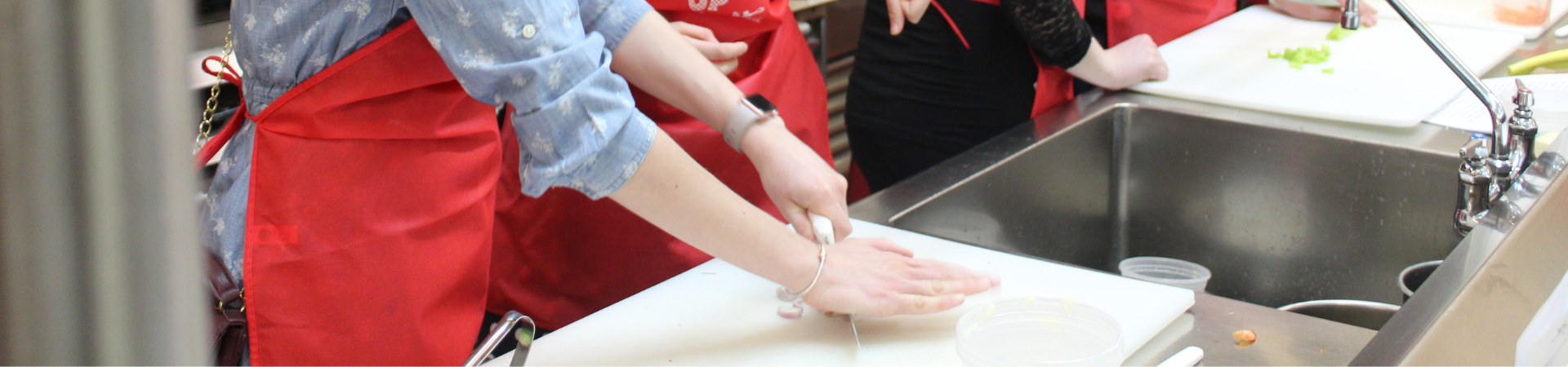 Individuals wearing red aprons are gathered around a kitchen counter, participating in a cooking activity. One person is chopping an onion on a white cutting board next to a sink.