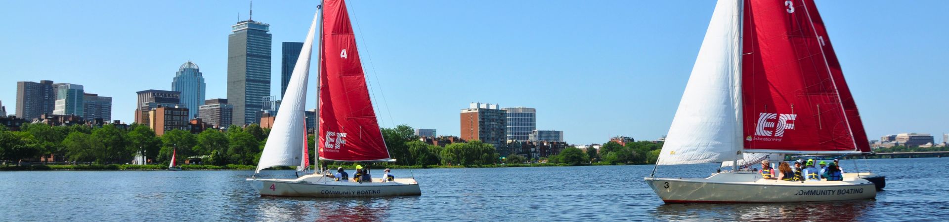 Two sailboats with red and white sails are on the Charles River with the Boston skyline in the background.