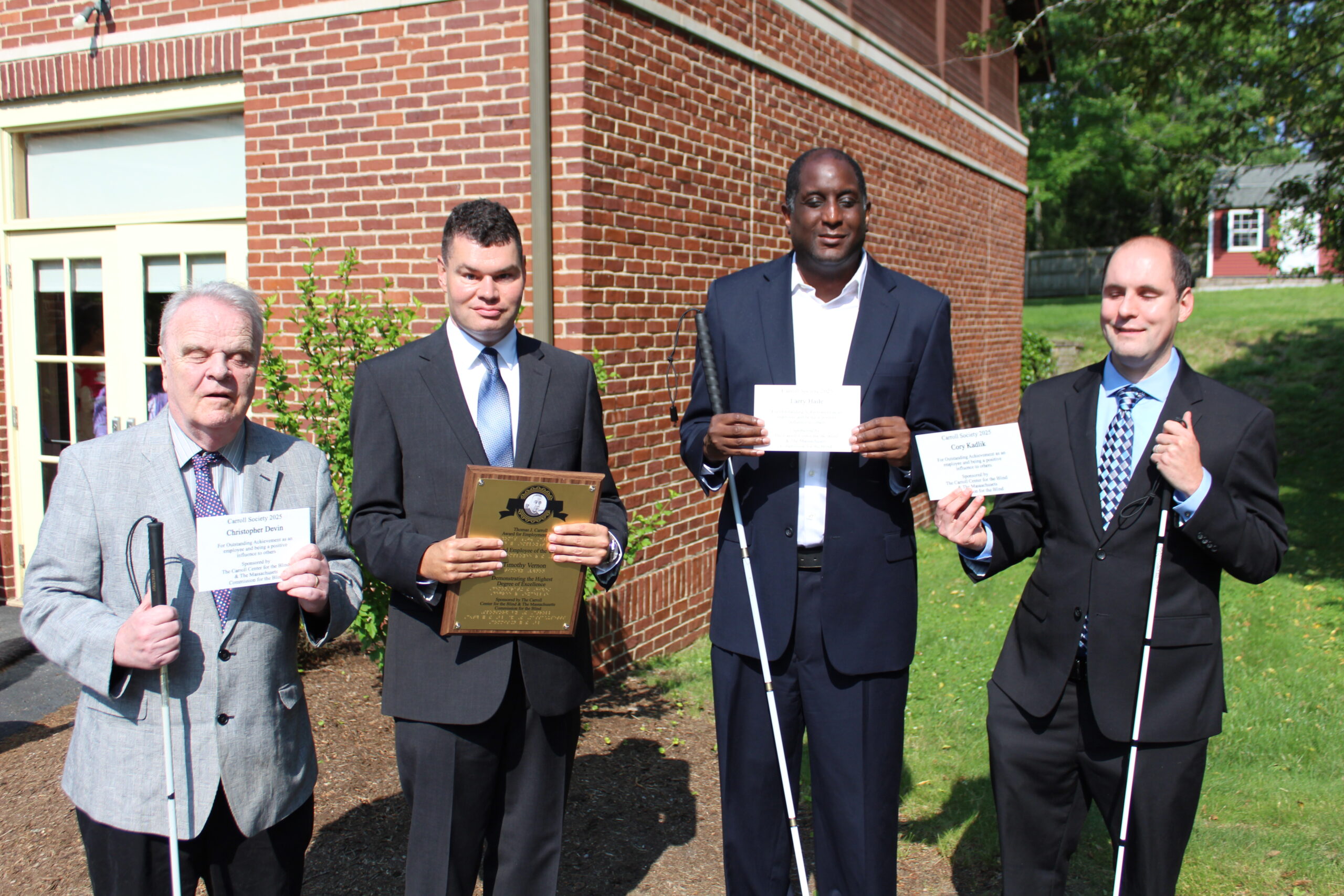 2025 Carroll Society Awards recipients stand outside a brick building, dressed in suits and holding awards. Each man is holding either a plaque or a certificate. All four men are using white canes, indicating they are blind or visually impaired. They are smiling and standing in a line, with greenery and a building in the background, on a sunny day.