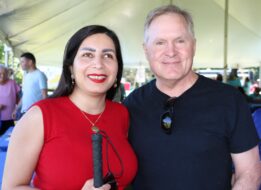Pardis is smiling while holding a white cane. She is wearing a red shirt with a gold necklace. Next to her there is a smiling man in a black T-shirt. They are outside underneath a big white tent.