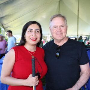 Pardis is smiling while holding a white cane. She is wearing a red shirt with a gold necklace. Next to her there is a smiling man in a black T-shirt. They are outside underneath a big white tent. 
