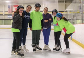 Ice Skate Six people stand together smiling, on the ice at an ice skating rink.