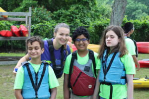 A photo of three Carroll Center summer youth program students stand with a summer program staff smiling and wearing life jackets outside. There are canoes and trees in the background.
