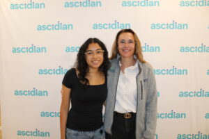 Real World of Work Experience Intern working at Ascidian. The photo shows the intern standing next to an employee at Ascidian with a backdrop that has the Ascidian logo