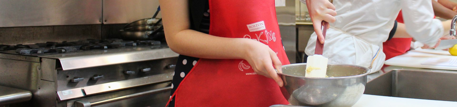 A person mixing ingredients in a bowl in a kitchen. The person is wearing a red apron and black t-shirt. There is a chef in a white apron and another baker in the back with a red apron.