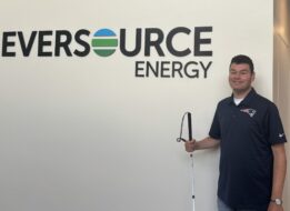 Tim Vernon with Eversource Logo Tim Vernon is standing next to the Eversource Energy logo on the wall while holding his white cane.
