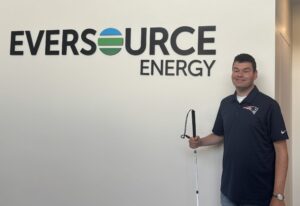 Tim Vernon is standing next to the Eversource Energy logo on the wall while holding his white cane.