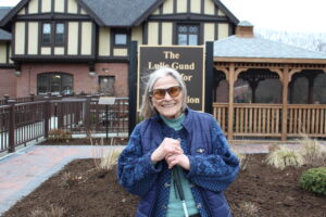 A senior client is smiling in front of the Lulie Gund Building. She is wearing a dark blue coat a turquoise shirt. She is also holding her white cane in front of her.