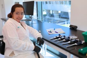 A smiling woman in a white lab coat and safety glasses sits at a laboratory chair with pipettes and scientific equipment, working in a modern research facility.