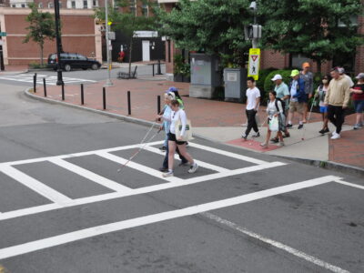 pairs of 2 crossing a cross walk. One individual is performing human guide for the blind sailor using a white cane.