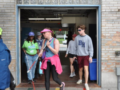 a group of blind sailors existing the lobby area of Community Boating and stepping onto the dock area. Volunteers are nearby to provide guidance.