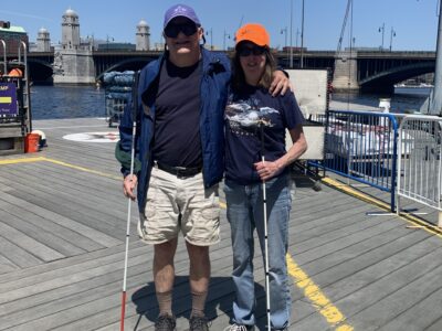 photo of 2 sailors (Michelle and Dave), standing next to each other on the docks. Dave has his arm around Michelle. Both are holding a white cane and wearing sneakers.