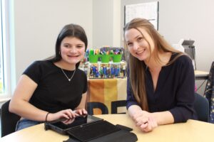 TVI and student Two young women sitting at a table, one using a tablet device, both smiling at the camera in an educational setting