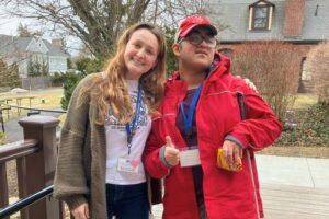 TVI and student volunteer TVI and Student Volunteer are smiling to the camera. They are outside and are both wearing Carroll Center lanyards. The TVI has long curly brown hair with a white shirt, brown cardigan, and black pants. The Student Volunteer has a red cap, red jacket, and is holding their cane and is givng a thumbs up.
