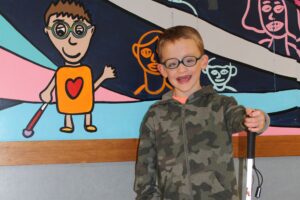 Ed Services Photo of kid A young smiling student is holding his cane and is wearing glasses and a sweatshirt. He stands next to a wall of paintings and one of them is of a boy with glasses, holding a cane, and is wearing a yellow shirt with a red heart.