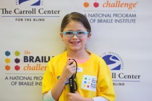 Ed Services Photo - Braille Challenge participant A Braille Challenge participant is smiling and holding there cane. They are standing in front of a Braille Challenge and Carroll Center banner.