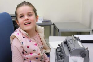 Ed Services Photo - Student Student is smiling and using a brailler. They are wearing a pink shirt and pink vest with rainbow hearts on it.
