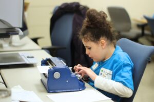 Ed Services Photo - Student on brailler Student is using a brailler at desk. They are wearing a blue Braille Challenge shirt.