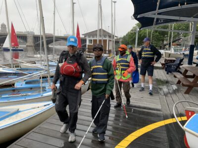 photo of a sighted guide walking with a blind sailor on the dock alongside boats tied up to the dock