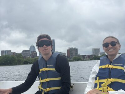 Two sighted guides in a sailboat (one male, Taylor, and one female, Morgan). Taylor is using the tiller to steer and wearing a blindfold for training purposes.