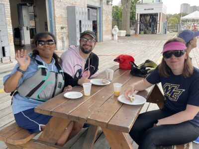 photo of 3 sailors sitting at a picnic table waving at the camera and smiling on the docks at Community Boating.