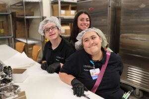Three smiling individuals, two wearing hairnets and gloves, sit at a work table in a kitchen.