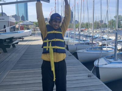 Sailor, Latha, raising her hands in the air in excitement, standing on the docks wearing a lifejacket.