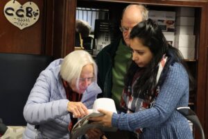 Low Vision Therapist demonstrates how to use a magnification device to an older woman in a light blue jacket and an older man in a green jacket. They're examining reading material together in the Carroll Store with dark wood paneling and a heart-shaped "CCB" sign with braille on it.