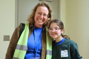 A woman with glasses and wavy hair wearing a brown jacket, blue shirt, and high-visibility vest stands posing with a young girl with glasses and a side ponytail. The girl is wearing a green long-sleeve shirt and glasses. Both are smiling at the camera in an indoor setting with beige walls.