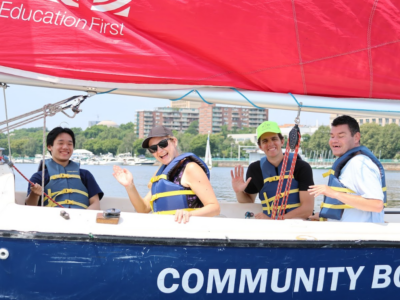 4 individuals sailing. All are smiling at the camera, two of them are waving "hello" at the camera.