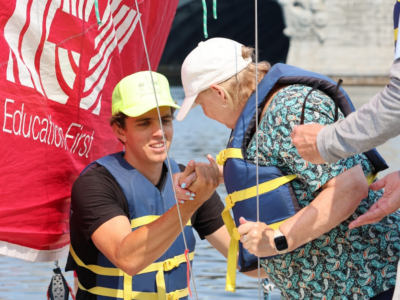 Photo of sighted guide, Taylor, assisting sailor, Laurie, safely step into the sailboat from the docks. Taylor is holding Laurie's arm.