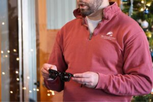 A man wearing a maroon quarter-zip pullover with "The Carroll Center for the Blind" logo holds a pair of meta AI glasses.
