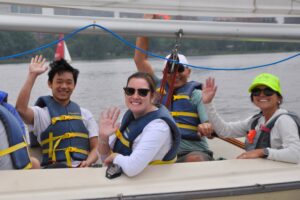 Four smiling people wearing life jackets wave from a sailboat on a river, with a city skyline visible in the background.
