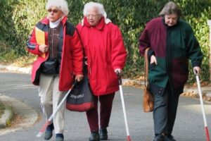 Three women walk side by side along a paved road or path, with green shrubs in the background. They use white mobility canes. The woman on the left wears a red and black jacket and carries a folder; the woman in the center wears a bright red coat and carries a bag; the woman on the right wears a dark green and maroon jacket and carries a purse.