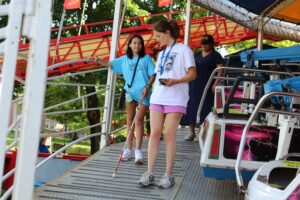 Mobility Training Photos (1) A young woman using a white cane navigates a metal ramp onto a carnival ride. A Summer Program A girl in a light blue t-shirt walks beside her, holding the railing. An adult in a dark blue dress stands in the background near the ride's cars. Red metal framework and green trees are visible in the background.
