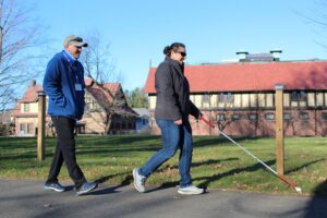 An Orientation & Mobility Instructor wallks behind a student using a white cane. The instructor is wearing a green cap and a blue jacket with a lanyard. The student wears sunglasses and a grey jacket and is holding her white cane.